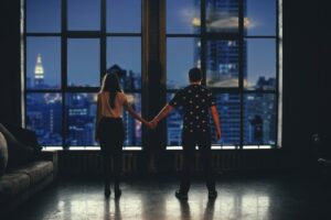 couple standing before a window looking over a city while holding hands.