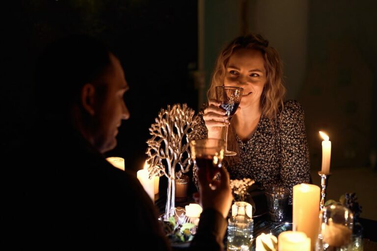 Couple on a date, focus is on woman who is raising a glass or wine. The lighting is dim.